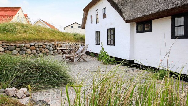A traditional white house with a thatched roof, stone wall, and a stone pathway leading to a wooden table and chairs. - Rustic Holiday Home in Løkken near Sea (Lokken)