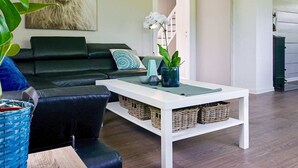 Ein modernes Wohnzimmer mit einem schwarzen Ledersofa, einem weißen Couchtisch und einem großen Gemälde von einem Schaf. Auf dem Couchtisch stehen Korbwannen und am Boden eine Topfpflanze.