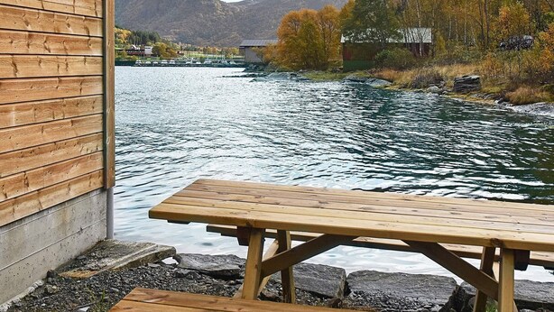 Eine Holzhütte mit Picknicktisch an einem See, umgeben von Herbstbäumen und Bergen.