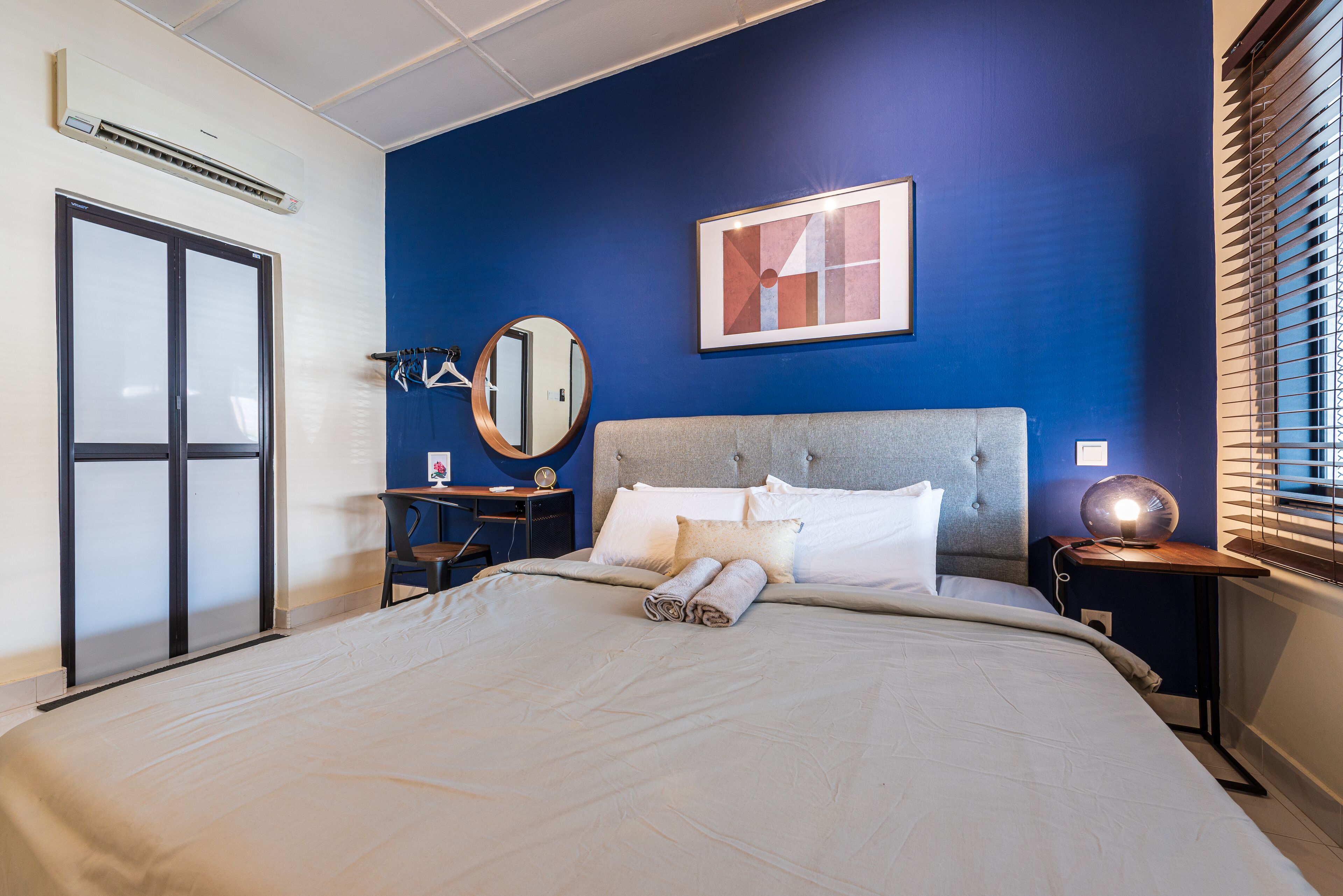 Ein Hotelzimmer mit einem großen Bett, einem Schreibtisch mit Stuhl, einem runden Spiegel, einem gerahmten Bild an der Wand und einem Fenster mit Jalousien.