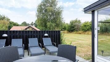 An outdoor seating area with a round table and four chairs, a wooden deck, a black fence, and a view of a grassy yard with trees.