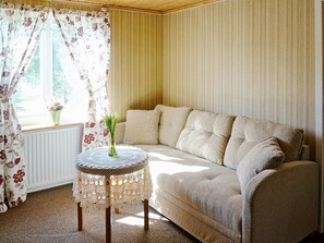 A cozy room with a beige sofa, a small round table with a lace tablecloth, and floral curtains. - Holiday Home in Ullared (Ullared)