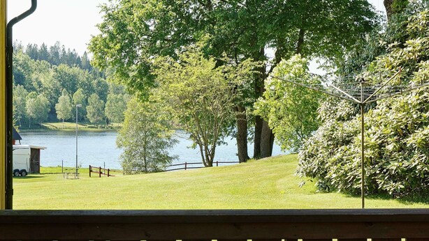 Une vue sur le lac depuis une terrasse en bois dotée d’une rambarde en métal, avec une zone herbeuse et des arbres.