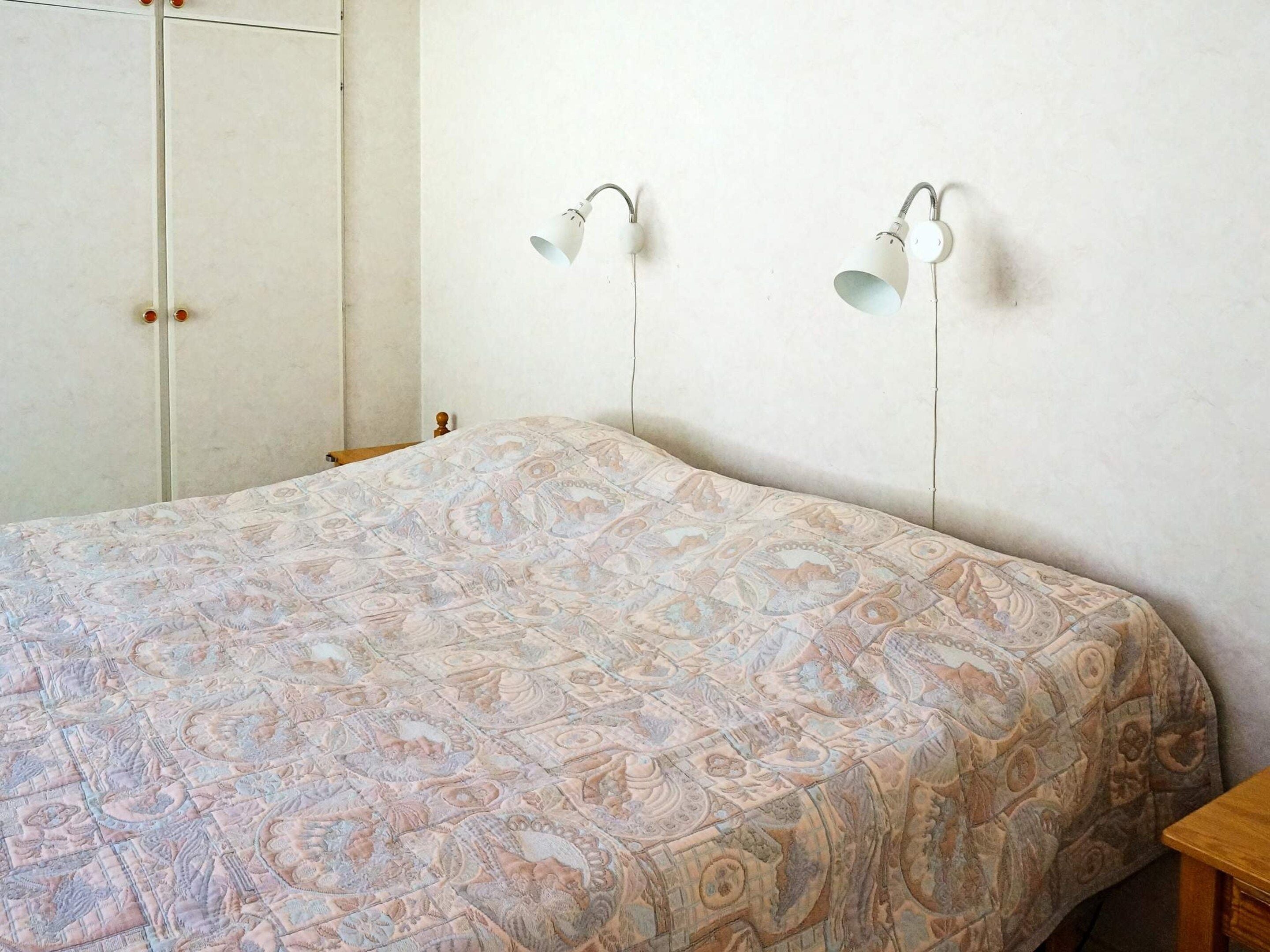 A bedroom with a patterned bedspread, a wooden headboard, and two wall-mounted lamps.