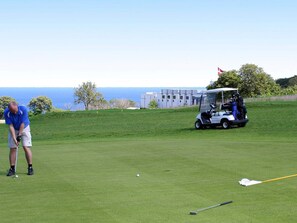 A golf course with a clear view of the sea in the background, a golf cart, and a golfer preparing to putt.
