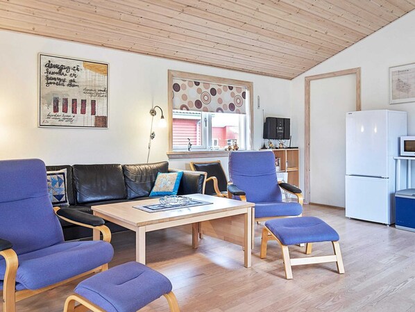 A modern living room with a wooden ceiling, a black leather sofa, blue armchairs, a wooden coffee table, and a white refrigerator. - 8 Person Holiday Home in Gudhjem (Gudhjem)