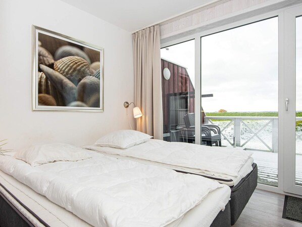 A bedroom with a bed, a nightstand, a wall-mounted lamp, and a large window overlooking a deck.
