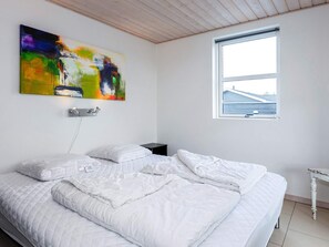 A bedroom with a bed, a colorful abstract painting, a nightstand, and a window with a view of buildings.