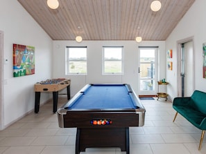 A pool table with a blue felt top, a foosball table, and a green sofa in a room with wooden ceiling beams and large windows. - 5 Star Holiday Home in Lokken (Lokken)
