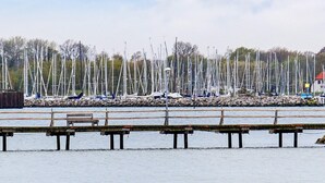 A marina with multiple sailboats docked, wooden benches along the water's edge, and a rocky shoreline. - Holiday Home in Gelting (Gelting)