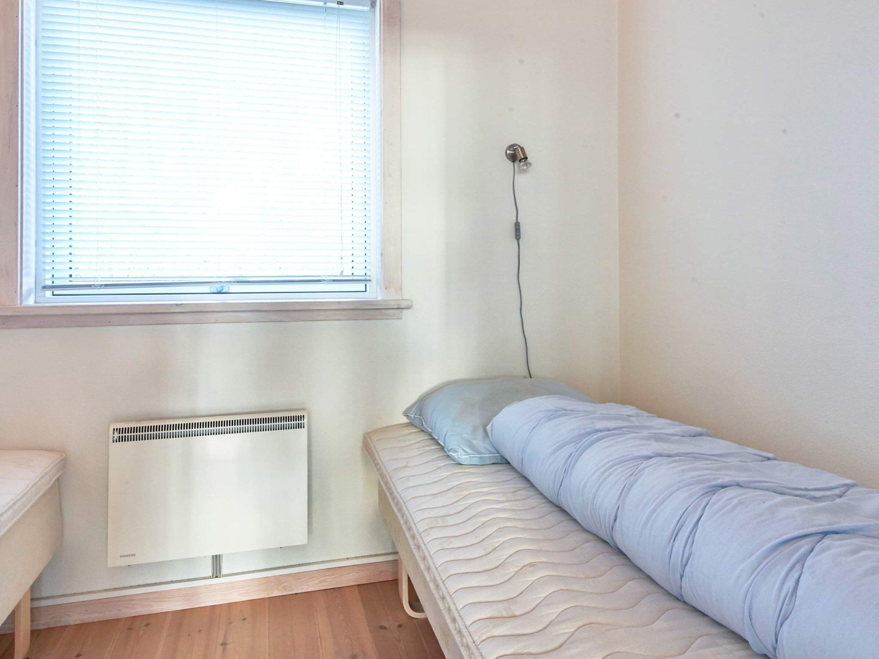 Un lit simple avec un matelas et un oreiller, un radiateur en dessous et une fenêtre avec des stores.
