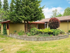 A single-story building with a red roof, a covered porch, and a garden area with various plants and a large tree. - 4 Person Holiday Home in Farvang (Farvang)