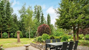 Une terrasse extérieure avec une table et des chaises, entourée d’un jardin où poussent différentes plantes et où se trouve une mangeoire à oiseaux.