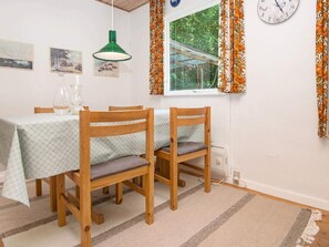 A dining area with two wooden chairs, a table with a patterned tablecloth, a green pendant light, and a window with floral curtains.