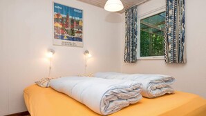 Una cama bien hecha con una colcha amarilla y sábanas blancas dobladas, un cartel de un pueblo y una ventana con cortinas estampadas.