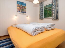 Una cama bien hecha con una colcha amarilla y sábanas blancas dobladas, un cartel de un pueblo y una ventana con cortinas estampadas.