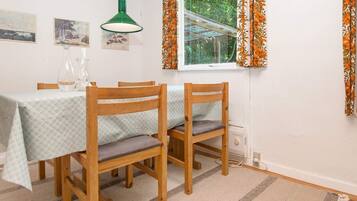 Un comedor con dos sillas de madera, una mesa con mantel estampado, una lámpara colgante verde y una ventana con cortinas anaranjadas.