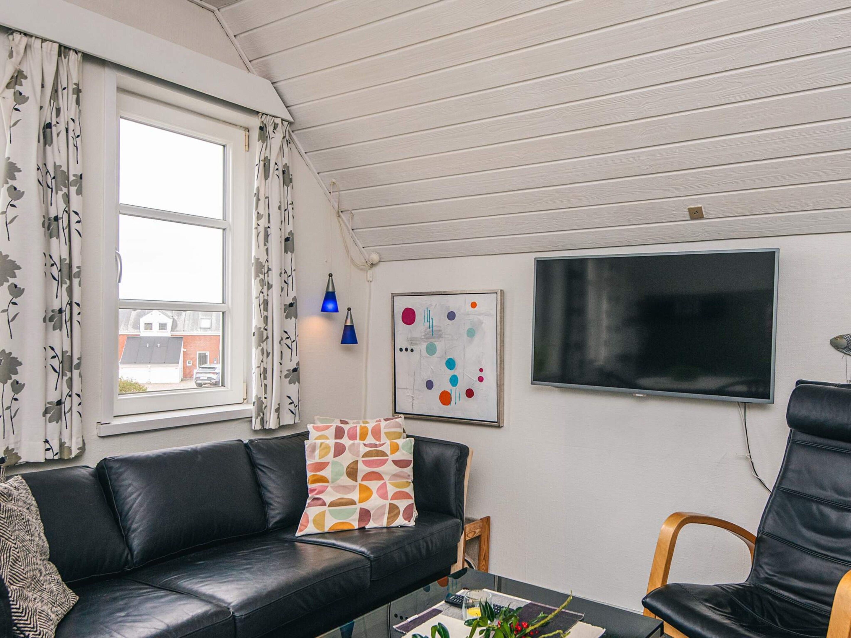 A modern living room with a black leather sofa, a wooden chair, a flat-screen TV, and a window with curtains.