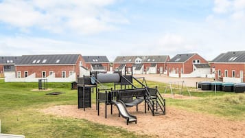 A playground with a slide and climbing structures, surrounded by a grassy area and red brick buildings with white trim.