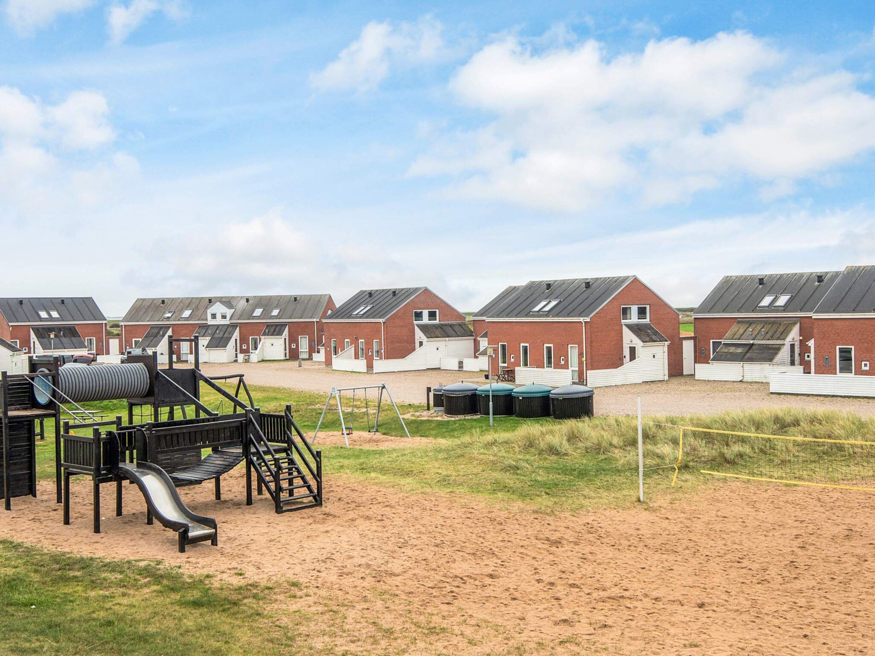 A row of red brick houses with white trim, a playground with a slide, and large black storage tanks in a sandy area.