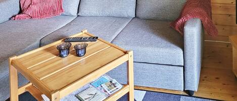A wooden coffee table with a magazine and remote controls, a grey sofa with a red blanket, and a wooden side table.