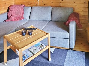 A wooden coffee table with a magazine and remote controls, a grey sofa with a red blanket, and a wooden side table. - 3 Person Holiday Home in Vastervik (Vastervik)