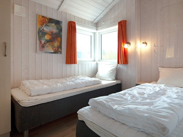 A room with two beds, a window with curtains, a painting on the wall, and a wall-mounted light fixture.