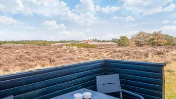 An outdoor seating area with a table and chairs, two cups, and a view of a field with trees and a red building in the distance.