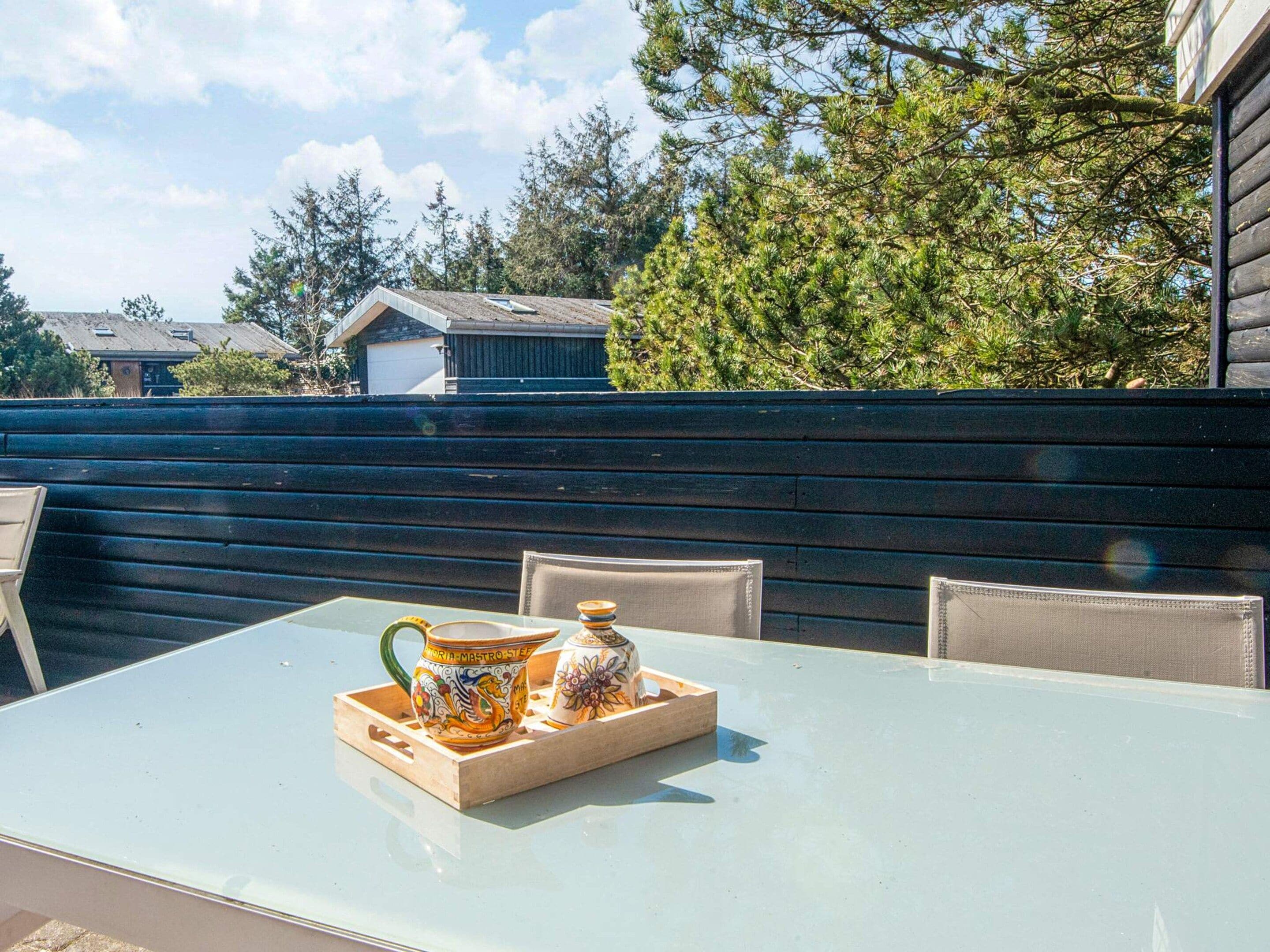 An outdoor patio with a glass table, two chairs, and ceramic containers on a wooden tray.