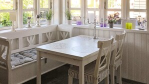 Uma área de refeições com mesa branca, quatro cadeiras, um banco com almofadas, e uma janela com plantas e um candelabro.