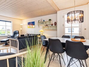 Un salon moderne avec un canapé noir, une table à manger avec des chaises, un lustre et une vue sur l’extérieur grâce à une grande fenêtre.