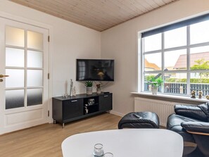 Ein modernes Wohnzimmer mit Fernseher, einem schwarzen Ledersofa, einem weißen Couchtisch und Blick ins Freie durch große Fenster.