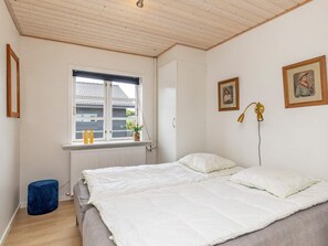 A bedroom with a bed, a window, a radiator, a blue ottoman, and two framed pictures on the wall.