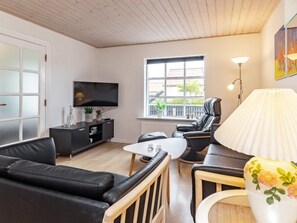 Un salon moderne avec un canapé en cuir noir, une table basse blanche, un fauteuil en cuir noir, un téléviseur posé sur un meuble noir et une fenêtre donnant sur l’extérieur.