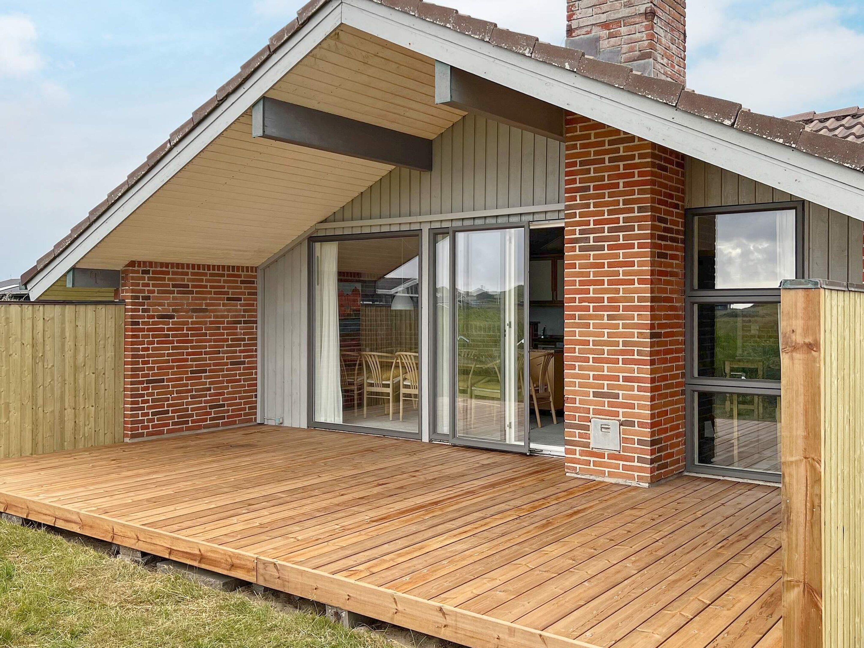 Una casa moderna con una terraza de madera, fachada de ladrillo y puerta de vidrio que da a una zona de estar al aire libre.