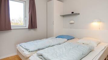 Ein Zimmer mit zwei Betten, einem fensterbelaubten Fenster, einem Wandregal und einer Wandlampe.
