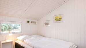 A single-bed room with white walls, a wooden ceiling, a bedside table with a lamp, and a window with a view of trees.