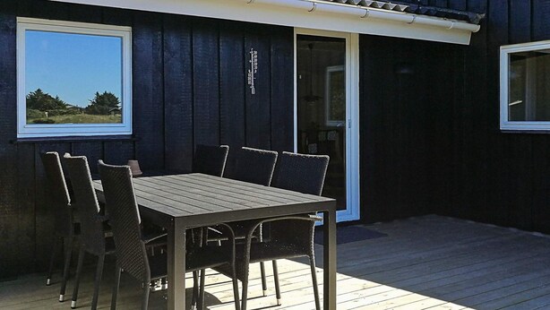 A covered outdoor patio with a table and chairs, a sliding glass door, and a window with a view of greenery.