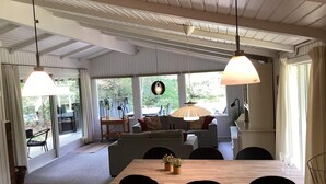 Un espace repas avec une table en bois, des chaises et des lampes suspendues. On aperçoit un canapé et une table à manger avec des chaises à l’arrière-plan.