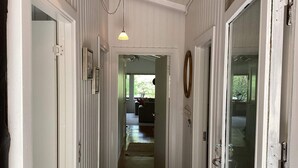 Un couloir étroit aux murs recouverts de panneaux blancs, un luminaire suspendu et une porte ornée d’un anneau en bois.