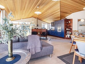 A modern interior with a wooden ceiling, a grey sofa, a dining table with chairs, and a kitchen area with blue cabinets.