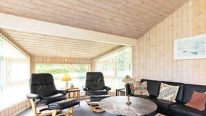 A cozy room with wooden panel walls, a black leather sofa, two leather chairs, a round coffee table, and a window with blinds. - 6 Person Holiday Home in Bindslev (Bindslev)