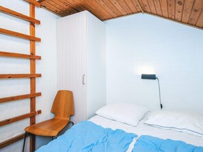 A small, single-room accommodation with a wooden ceiling, a bed with white bedding, a chair, and a wardrobe.