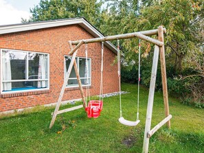 A wooden swing set in front of a brick house with a window and a red bucket.
