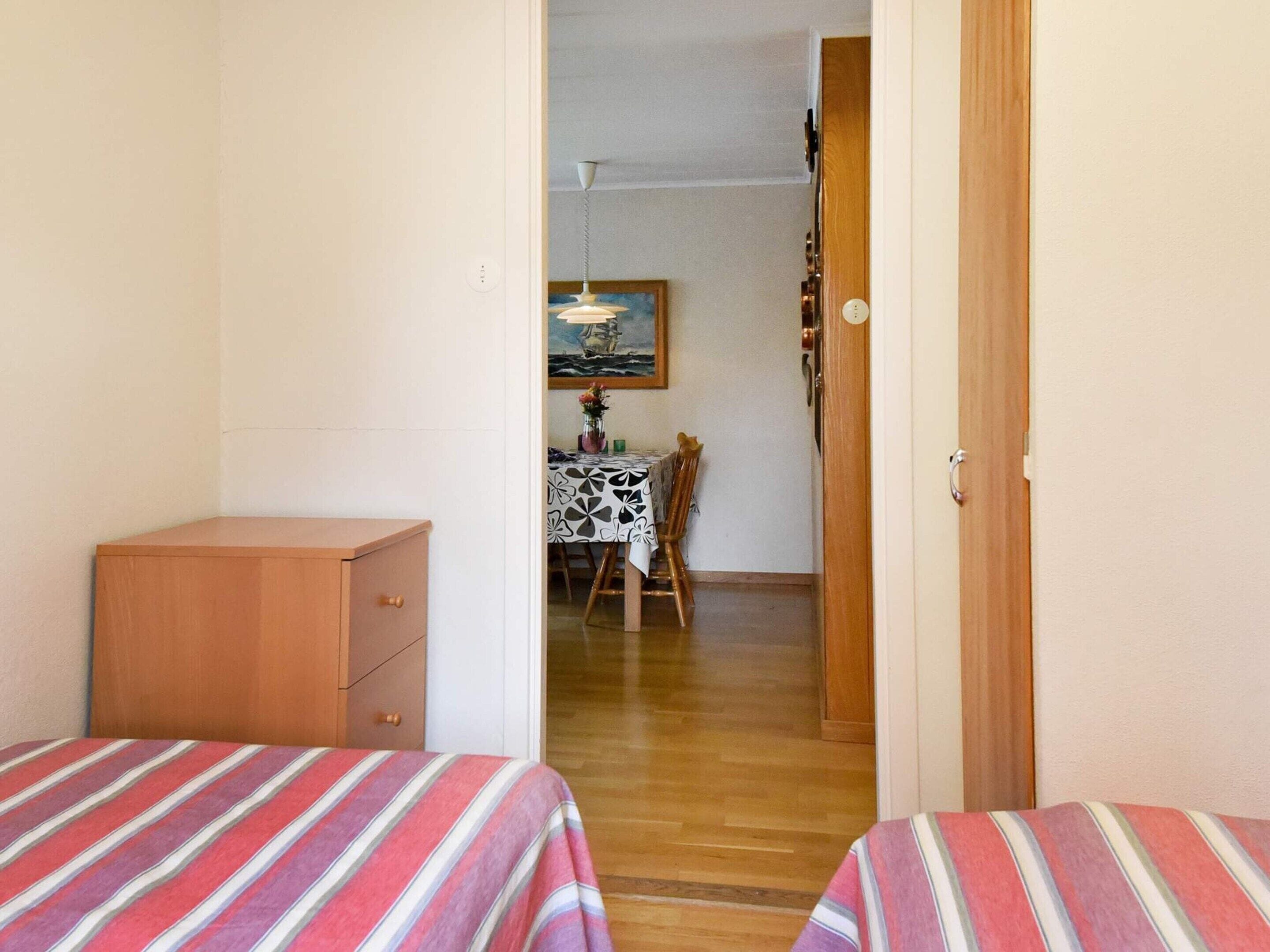 A room with two beds, a wooden dresser, a dining table with a patterned tablecloth, and a painting on the wall.