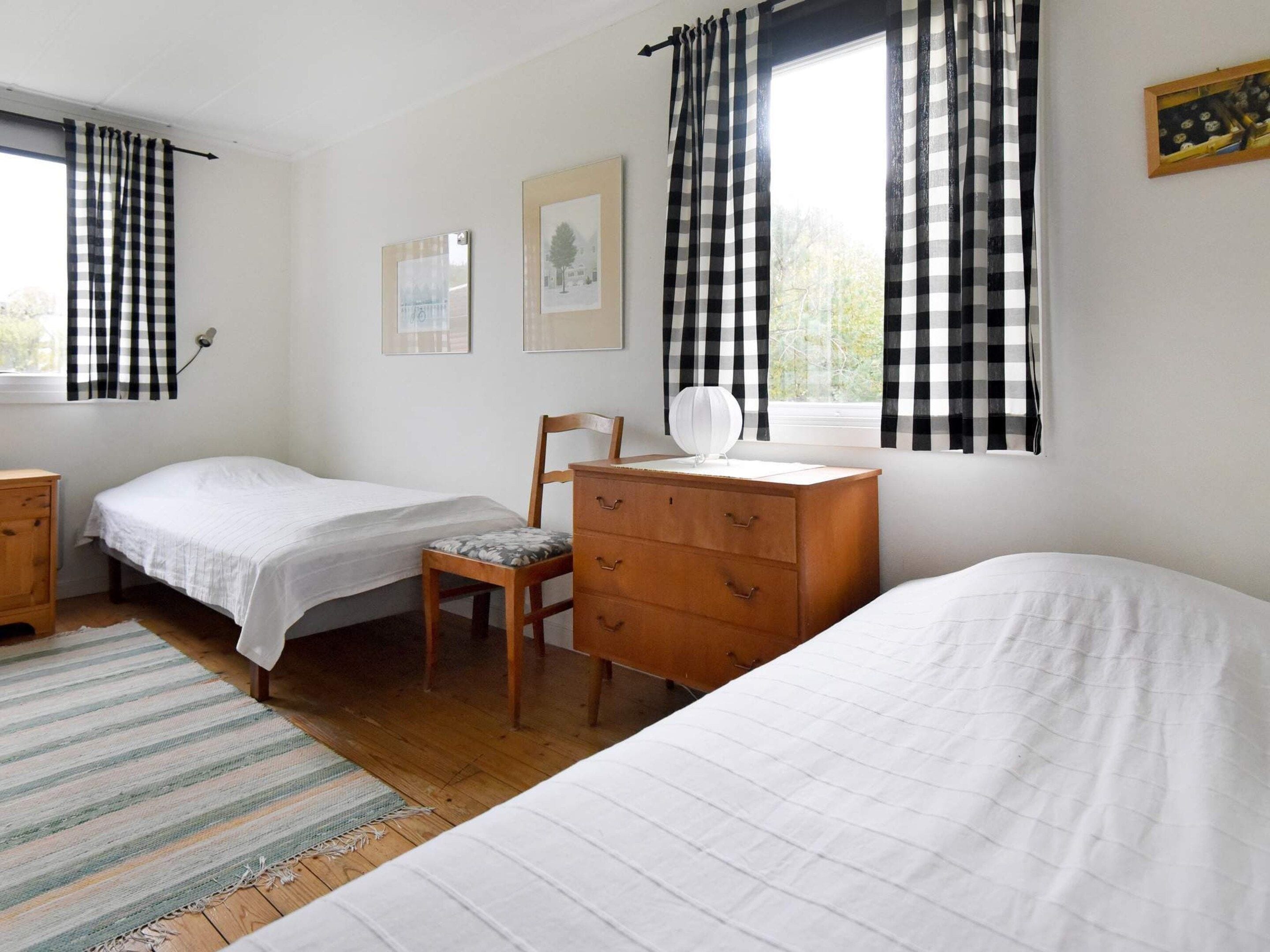 A bedroom with two single beds, a wooden dresser, a chair, a window with checkered curtains, and framed pictures on the wall.
