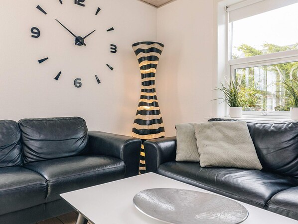 A modern living room with a black leather sofa, a white coffee table, a large wall clock, a tall striped vase, and a window with plants.