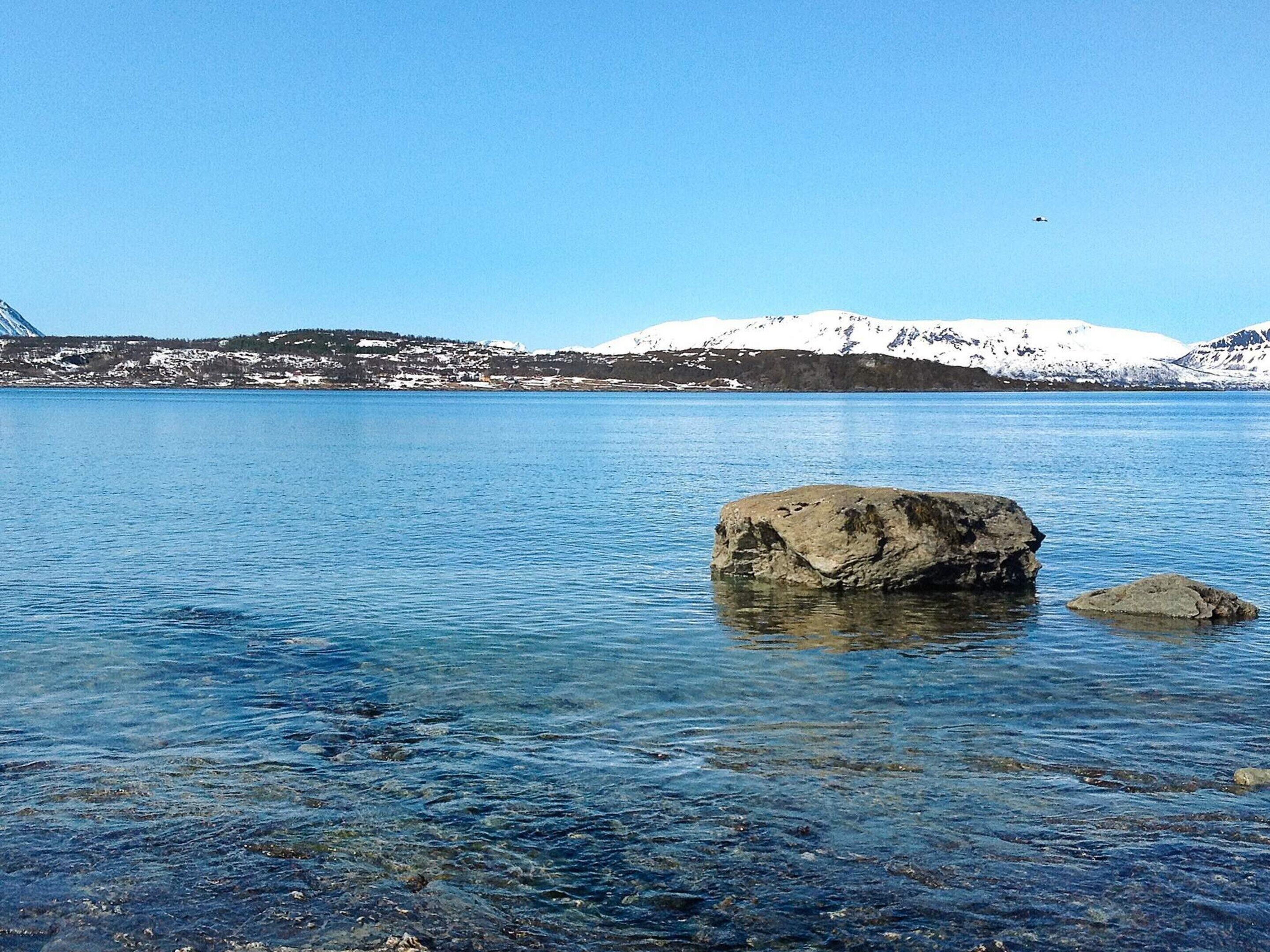 A clear lake with a large rock in the foreground and snow-capped mountains in the background.