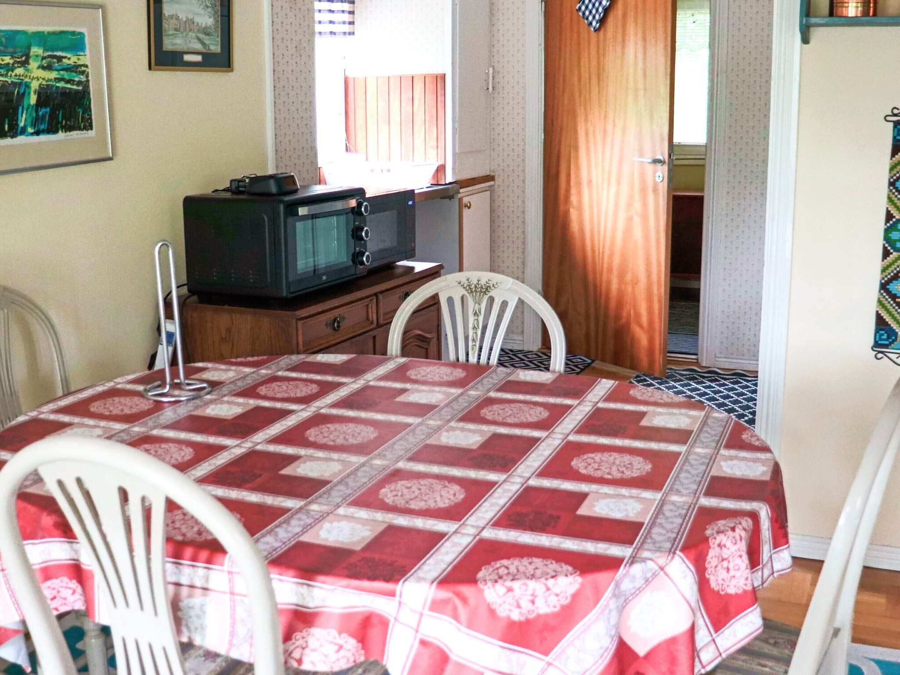 A dining area with a table covered in a red and white patterned tablecloth, white chairs, a wooden cabinet with a television, and a window with curtains.