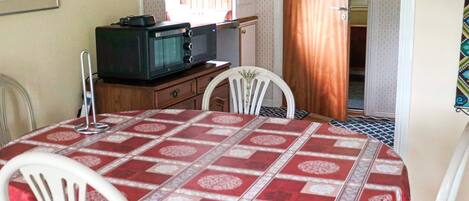 A dining area with a table covered in a red and white patterned tablecloth, white chairs, a wooden cabinet with a television, and a window with curtains.
